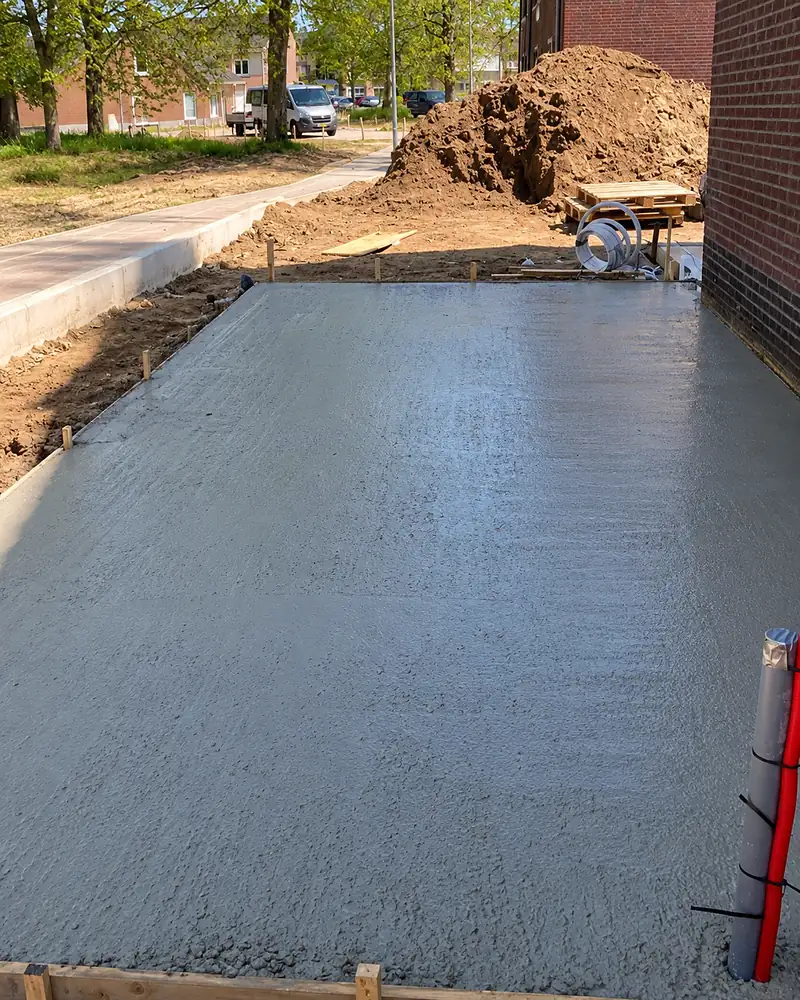 Pas gestorte betonvloer met een geborstelde afwerking naast een woning in aanbouw.