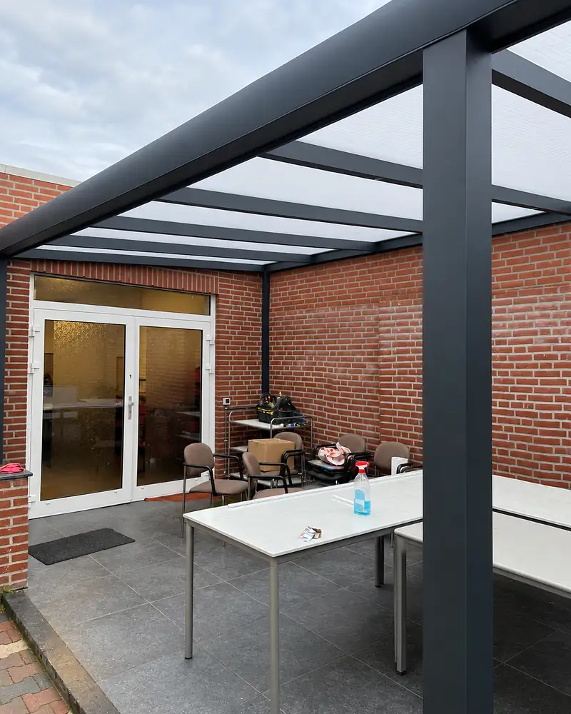 Afgewerkte antracietkleurige overkapping met lichtdoorlatend dak tegen een gebouw van rode baksteen.