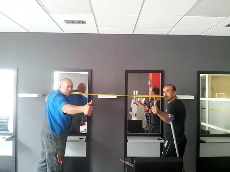 Twee vakmensen steken hun duim op in een net opgeleverde kapsalon met donkergrijze wanden en spiegels.