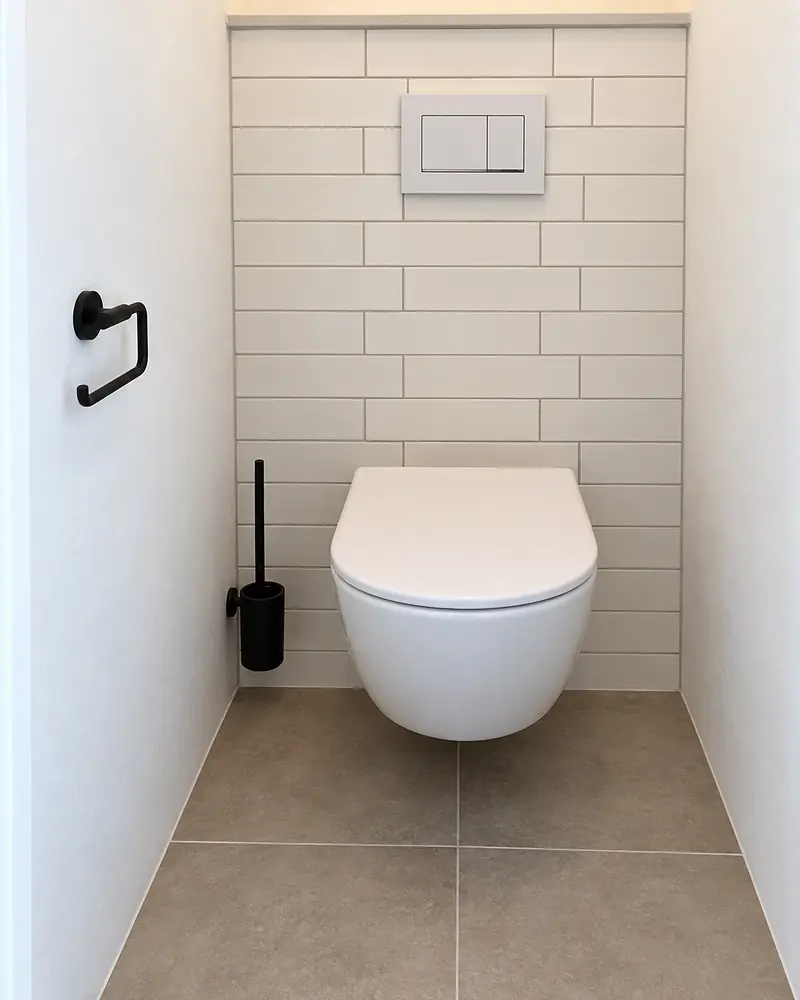 Afgewerkt toilet met witte wandtegels in halfsteensverband, een hangend toilet en grijze vloertegels.